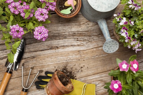 The image depicts a wooden outdoor garden table with a weathered texture, featuring various gardening tools and potted plants. On the table, there are pink and purple flowering plants, a small terracotta pot filled with soil, and a yellow gardening glove. A metal hand rake is placed beside the pot, along with a small trowel with a wooden handle. A grey watering can with a round spout and a drainage hole is positioned on the table, suggesting ongoing garden maintenance. The scene is set outdoors, in natural daylight, possibly during a mild weather day suitable for garden work. The arrangement indicates a typical gardening activity setup, and the presence of the gloves and tools suggests routine lawn and plant care. Such an image aligns with the services provided by Gardening Services Finsbury Park, emphasizing general garden upkeep, planting, and outdoor space maintenance in the local area, with a focus on vibrant plant life and outdoor horticultural tasks. The description is neutral, professional, and aimed at supporting accessibility and local SEO relevance for gardening and landscaping services in Finsbury Park postcode area.