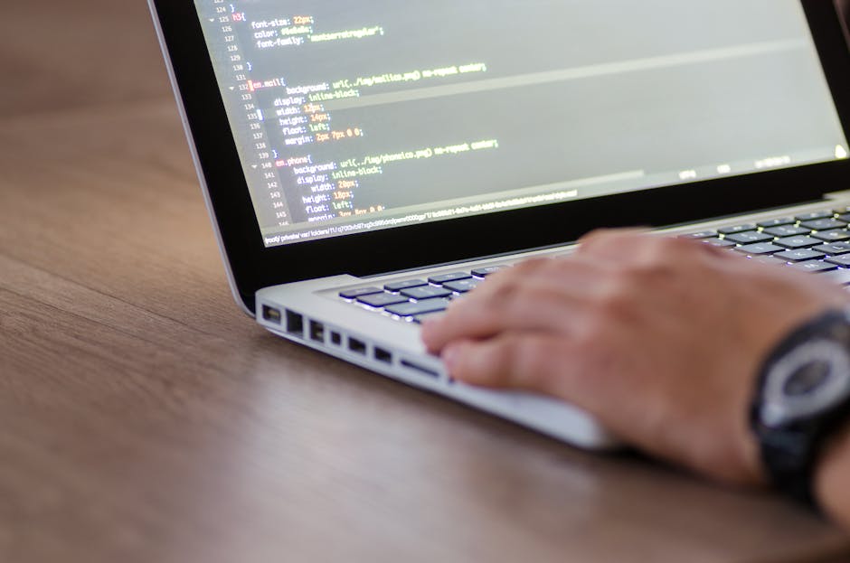 A person operating a laptop on a wooden desk, with the screen displaying lines of computer code in a text editor. The individual's hand is resting on the keyboard, with a black wristwatch visible on t
